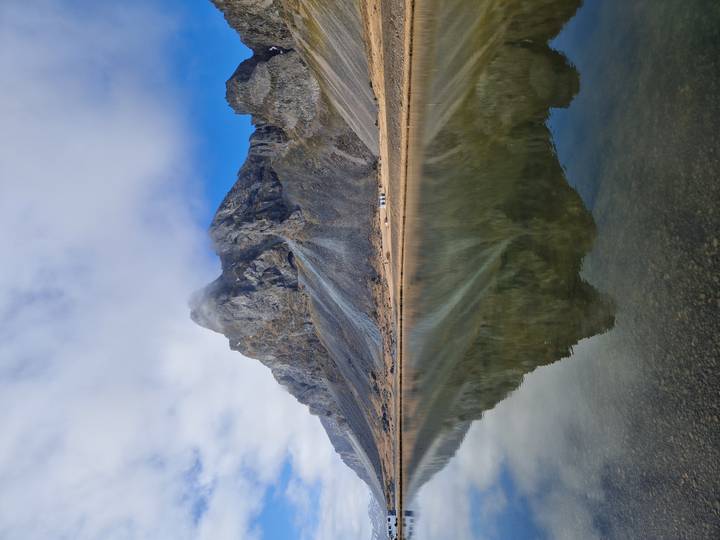 Craggy mountain reflected perfectly in calm lake water beneath patchy clouds and blue sky.