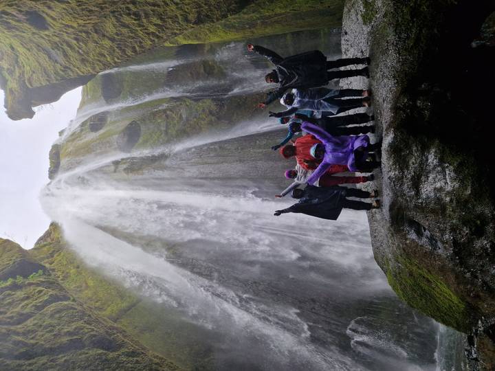 Rain-clad group raises arms in triumph beneath a thundering waterfall inside a rocky cavern.