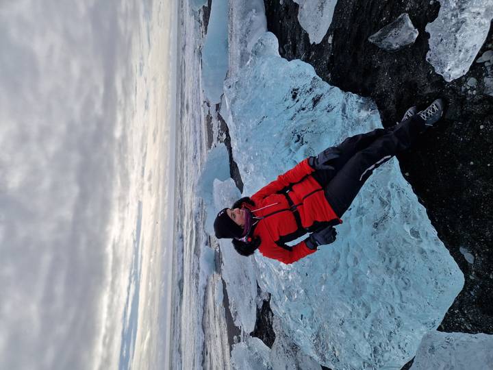 Woman in red jacket lounges on a blue iceberg chunk along a black-sand beach under grey skies.