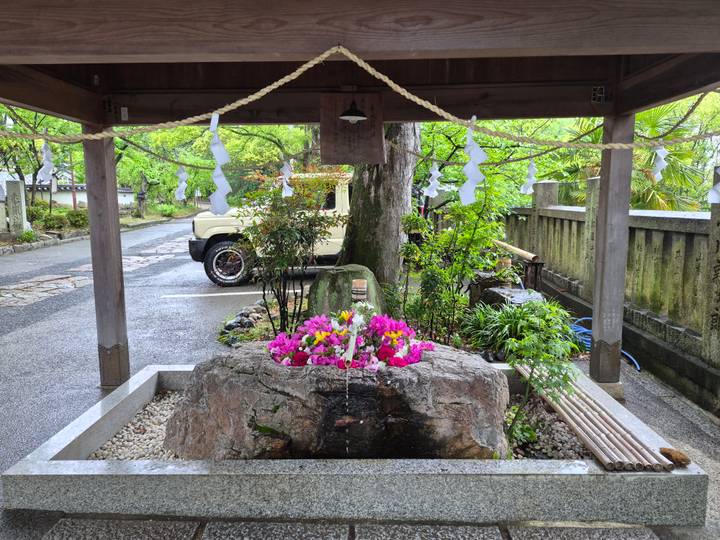 Stone purification fountain decorated with fresh flowers beneath a wooden Shinto shrine roof and sacred rope.
