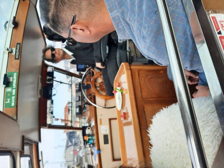 Captain steers a small river boat while passengers observe from the cabin.