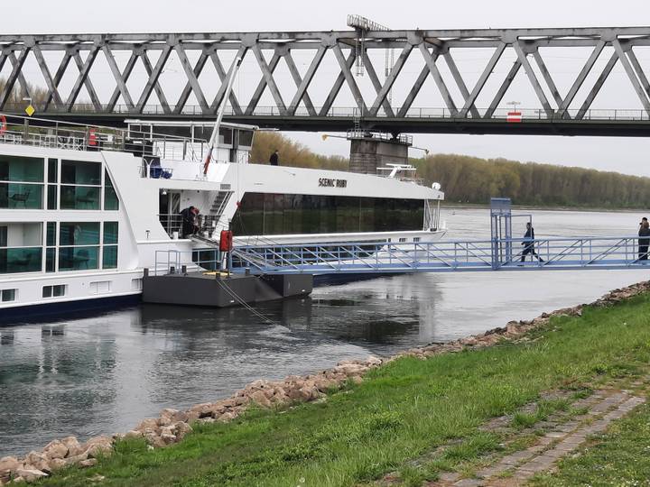 Modern white river-cruise vessel moored beside a bridge, with passenger walking the gangway.