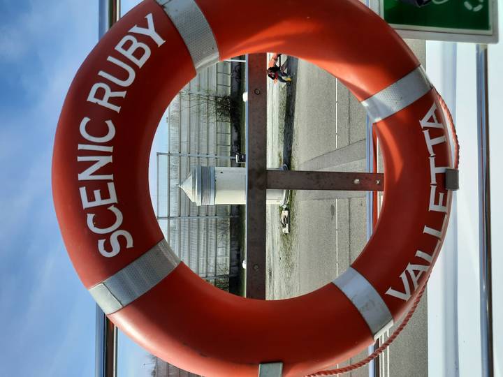 Close-up of orange life ring labeled 'Scenic Ruby' aboard a ship deck under blue sky.