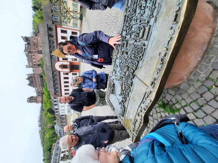 Tour guide explains a bronze tactile model of Heidelberg with castle ruins on hill behind.