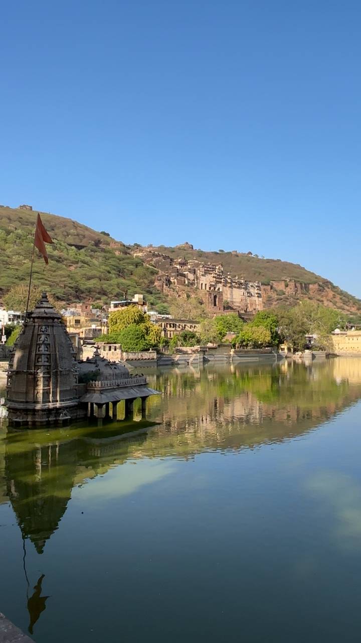 Historic Bundi palace and hillside fort are reflected in a tranquil lake beneath a clear blue sky.
