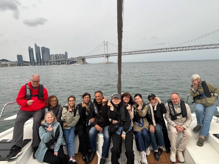 Tour group in lifejackets poses on boat deck with Busan skyline and suspension bridge behind.