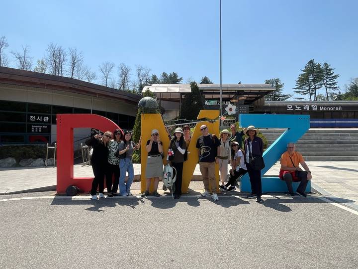 Travelers cheerfully gather before colorful DMZ sign outside visitor center on sunny day.