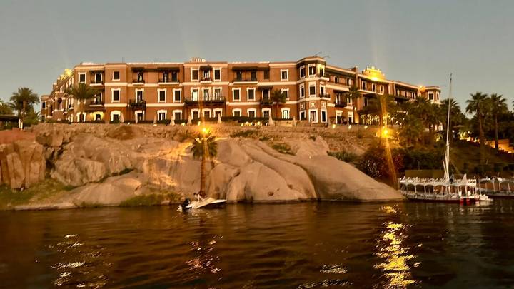 Historic riverside hotel atop granite rocks illuminated at dusk along the Nile.