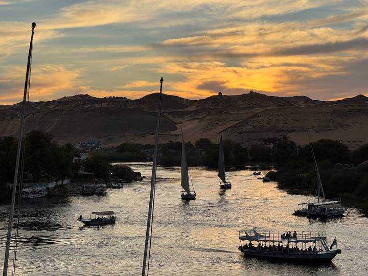 Feluccas sail the Nile at sunset with dramatic orange clouds over desert hills.
