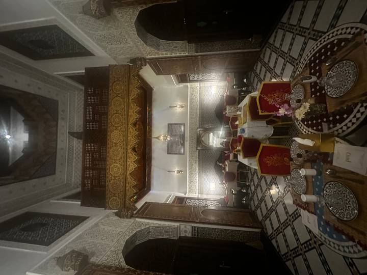 Dimly lit traditional riad dining room with patterned tiles and ornate wooden balcony.