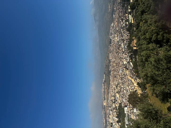 Wide panoramic view over a sprawling Moroccan city nestled between green hills under a clear blue sky.