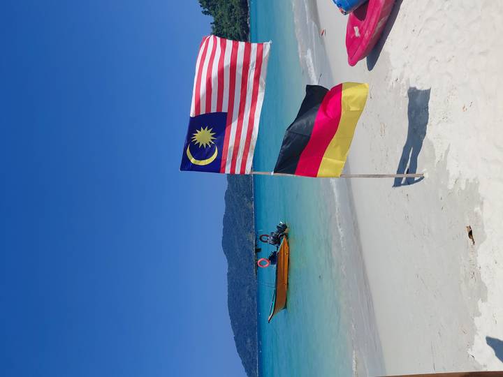 Pristine white-sand beach with Malaysian and German flags waving beside a small wooden boat on turquoise water.