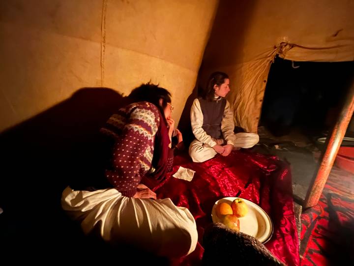 Dimly lit interior of a desert tent with two travelers sitting on red carpets beside a plate of fruit.