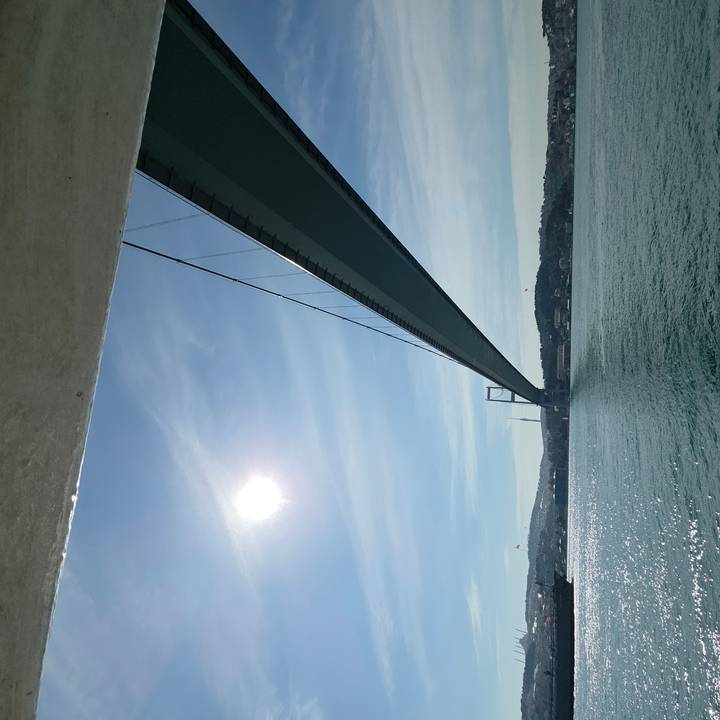A long suspension bridge stretches across the Bosphorus with bright sun and blue sky overhead.
