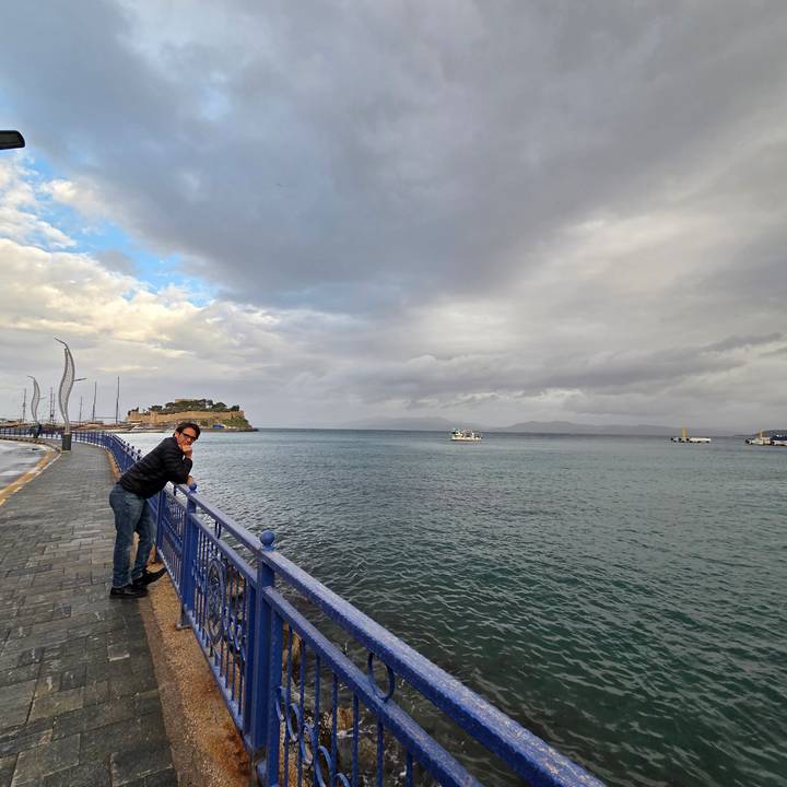 A traveler leans on a blue seaside railing, looking out over a bay with distant boats and a cloudy sky.
