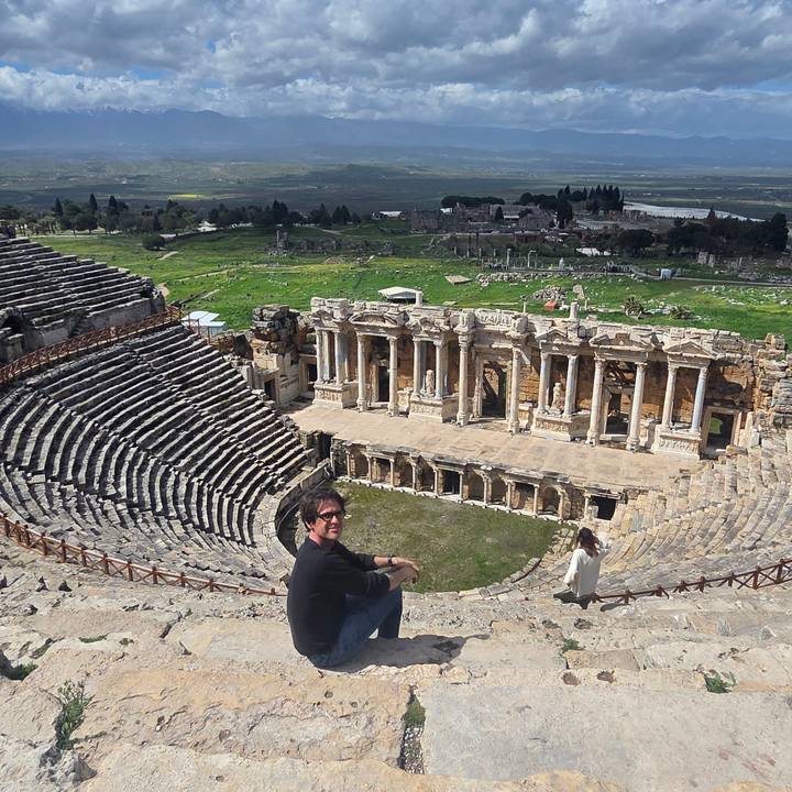 Travelers sit on the ancient stone tiers of Hierapolis’s Roman theatre overlooking its ornate stage façade and surrounding green plain.