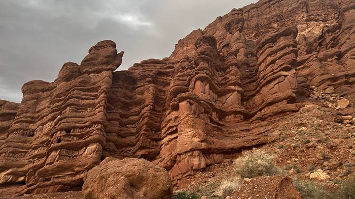 Towering layered red rock cliffs rise toward a cloudy sky in a rugged canyon landscape.