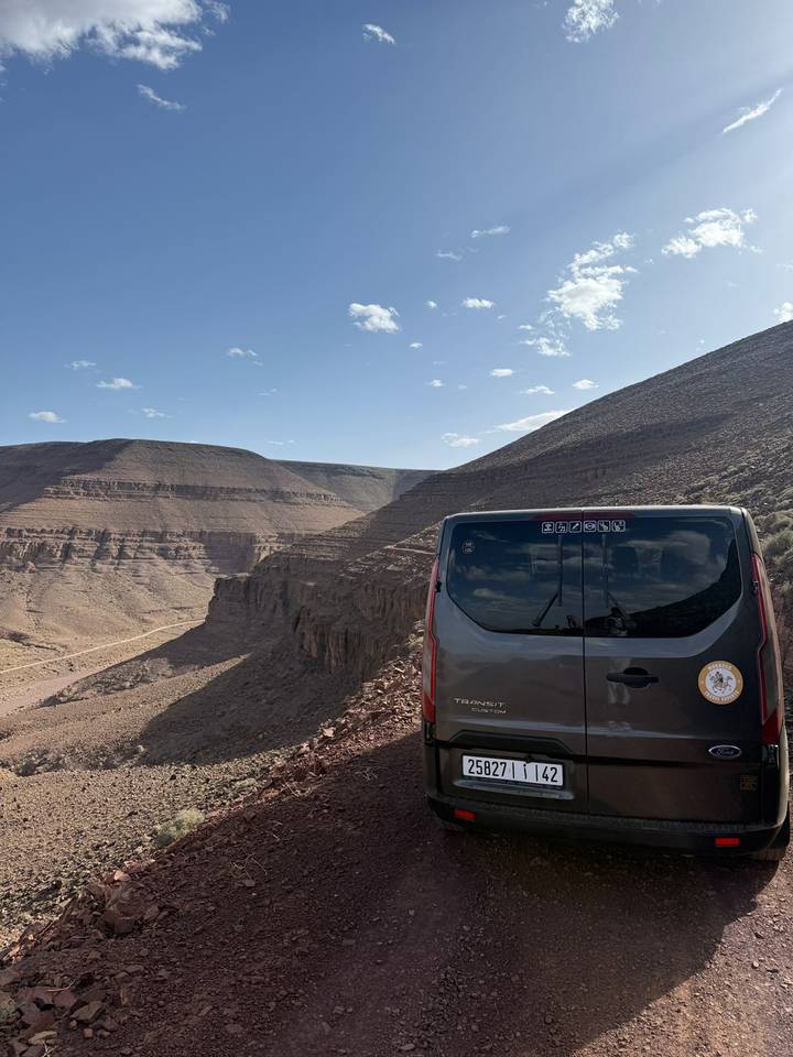 A tour van parks at a high desert overlook with steep eroded cliffs and a winding dirt road below.