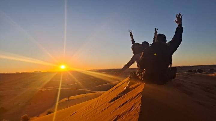 Two travelers sit atop a golden sand dune raising their arms as the sun sets over the Sahara horizon.