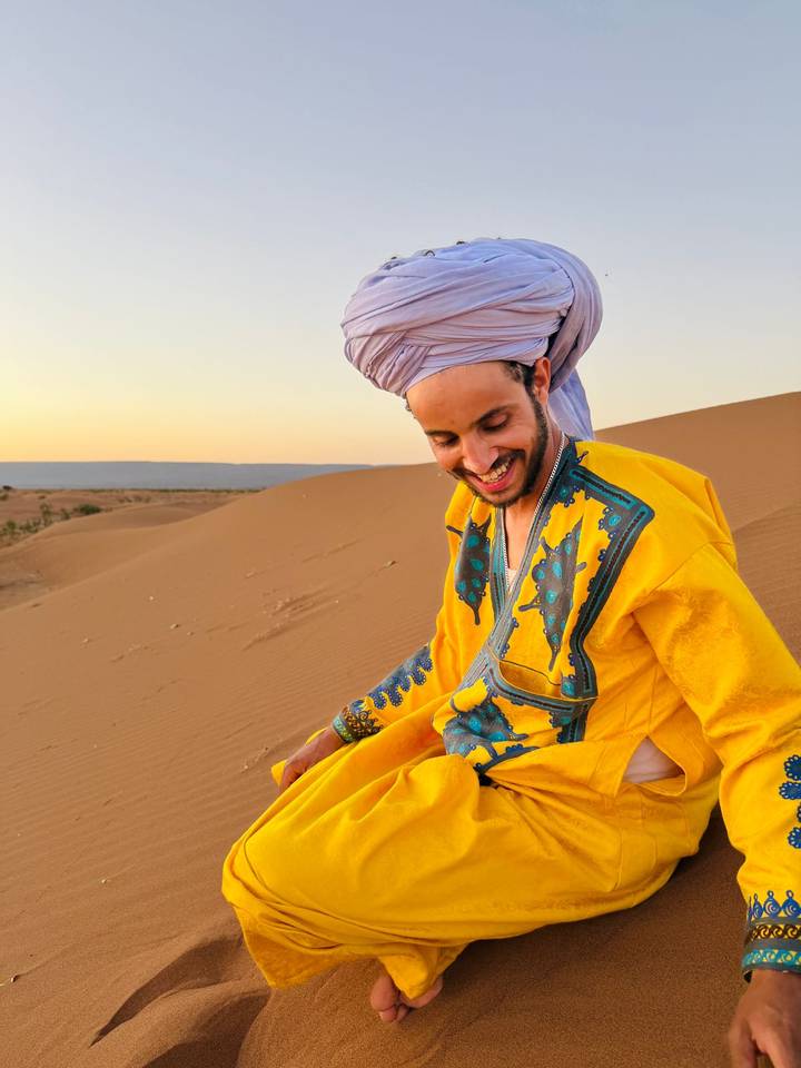 A smiling man in bright traditional clothing and turban sits on warm desert sand at golden hour.