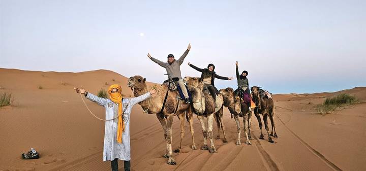 A caravan of tourists rides camels across soft desert sand under a pale sky with the moon visible.
