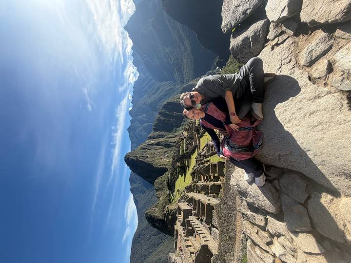 Couple seated on a rock ledge overlooking the terraces and peaks of Machu Picchu with clear blue sky