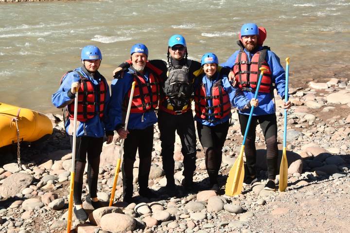 Five rafters in helmets and life jackets pose with paddles beside a fast-flowing river