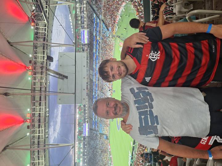 Father and son pose inside a lively football stadium with crowd and pitch behind them