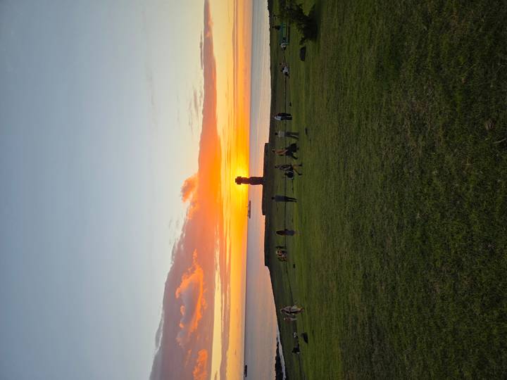 Visitors watch a vivid orange sunset behind a solitary Moai near the ocean shoreline