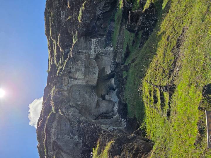 Rocky cave entrance cut into cliff face with grassy foreground steps and sun overhead