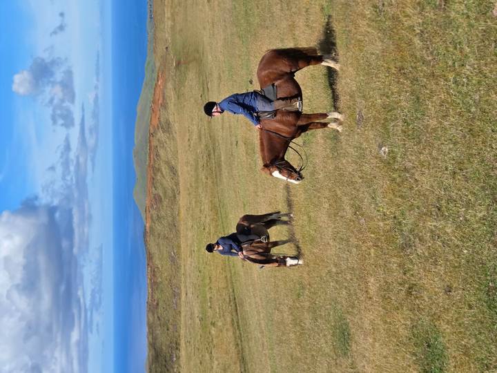 Two riders on horseback traverse green rolling hills with blue ocean horizon beyond