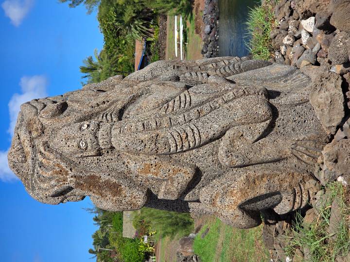 Large weathered stone carving featuring stylised human and animal forms beside a small lagoon