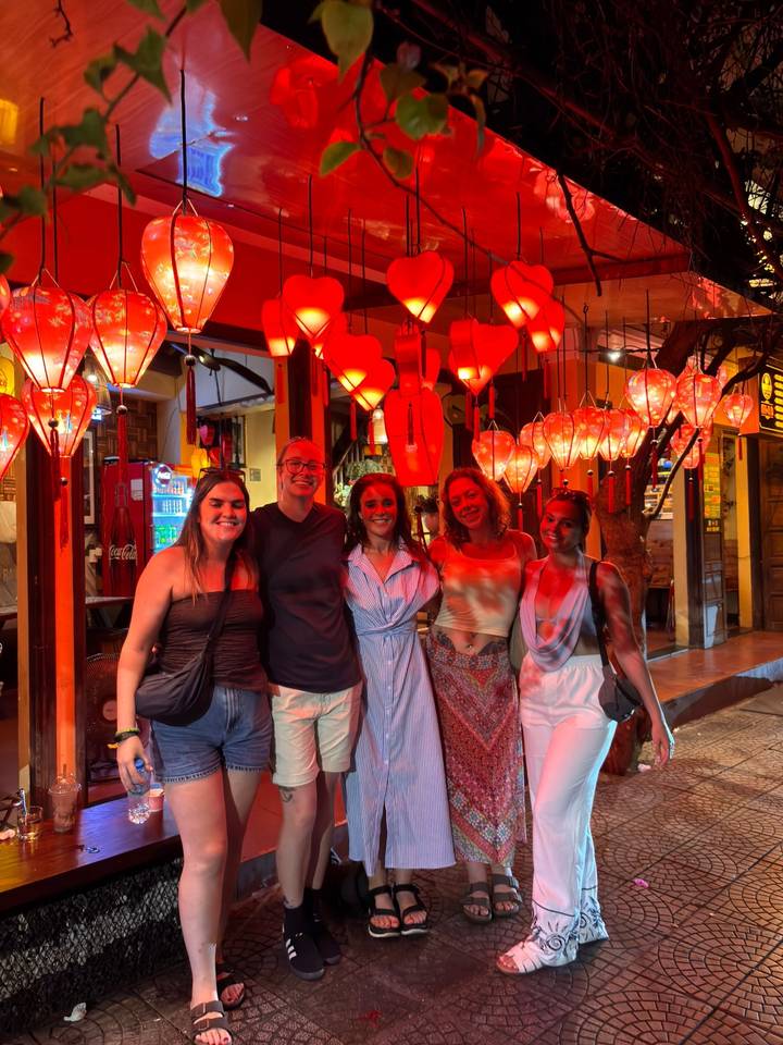 Five young women smiling beneath rows of glowing red lanterns in a vibrant street setting.