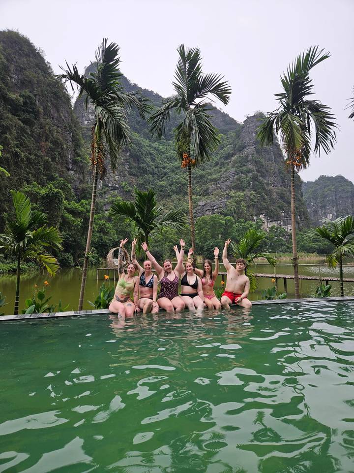 Group of friends in swimwear cheering from an infinity pool with limestone cliffs and lush forest behind.