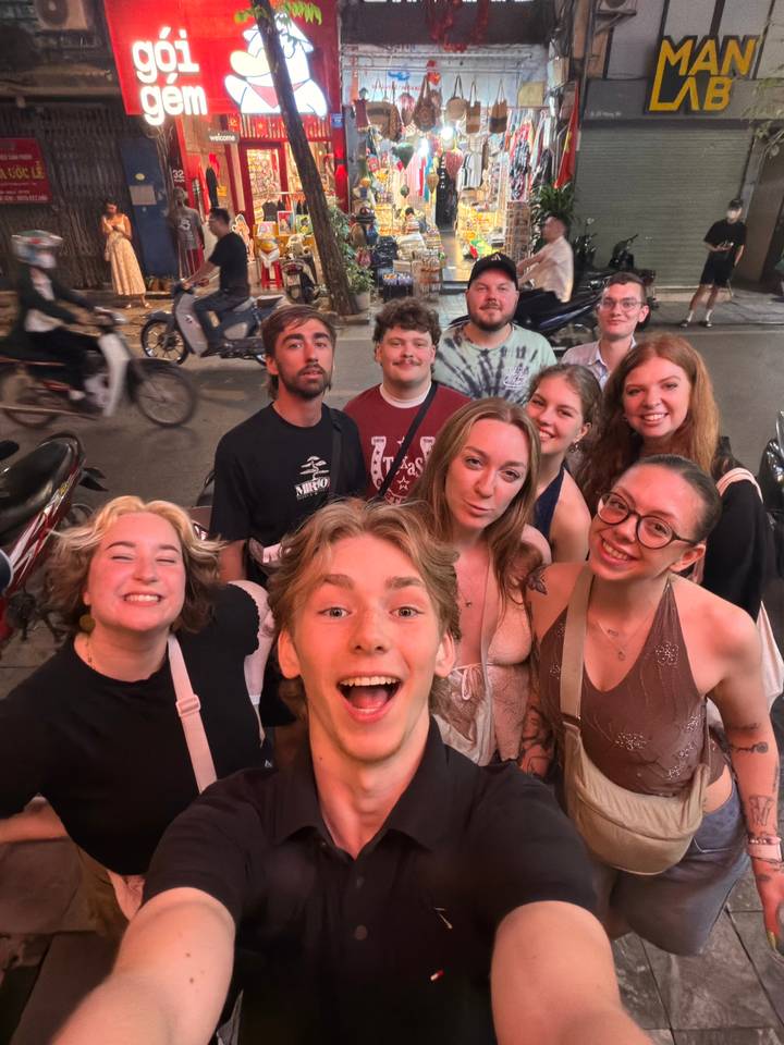 Large group selfie on a lively Vietnamese street with scooters and neon lights around.