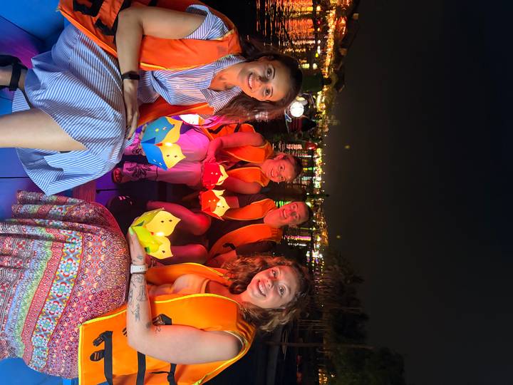 Four women in life jackets holding colorful paper lanterns while sitting in a small boat on a river at night.