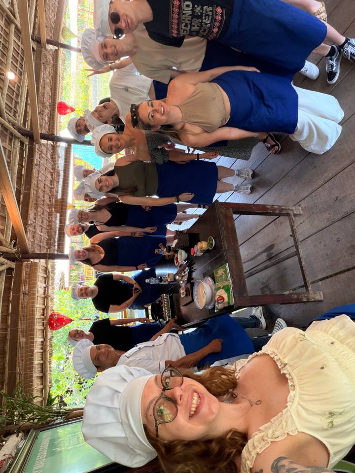 Cooking class participants wearing blue aprons and chef hats around a prep table with local ingredients.