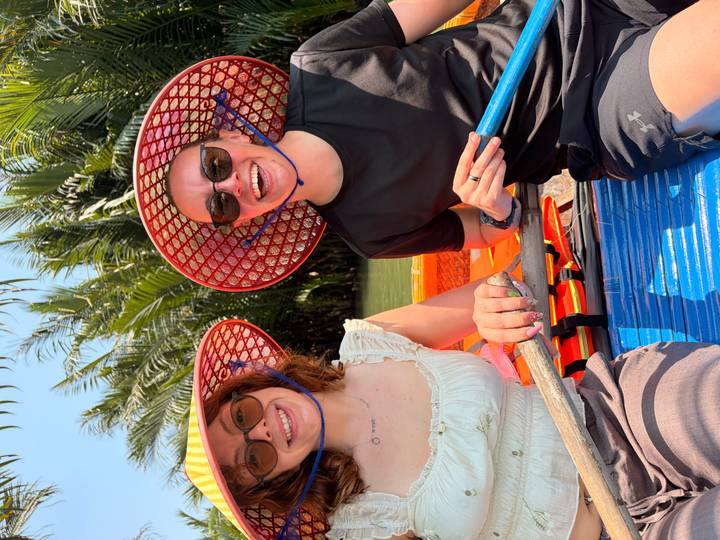 Two travellers in conical hats smiling while rowing a small boat through lush palm-lined waters.