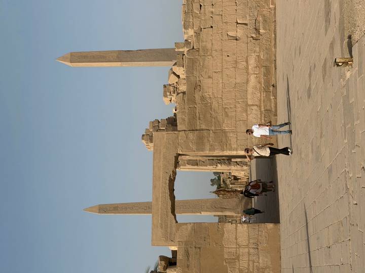 Visitors walk through a sandstone gateway flanked by towering obelisks and ruined temple walls under a clear blue sky.