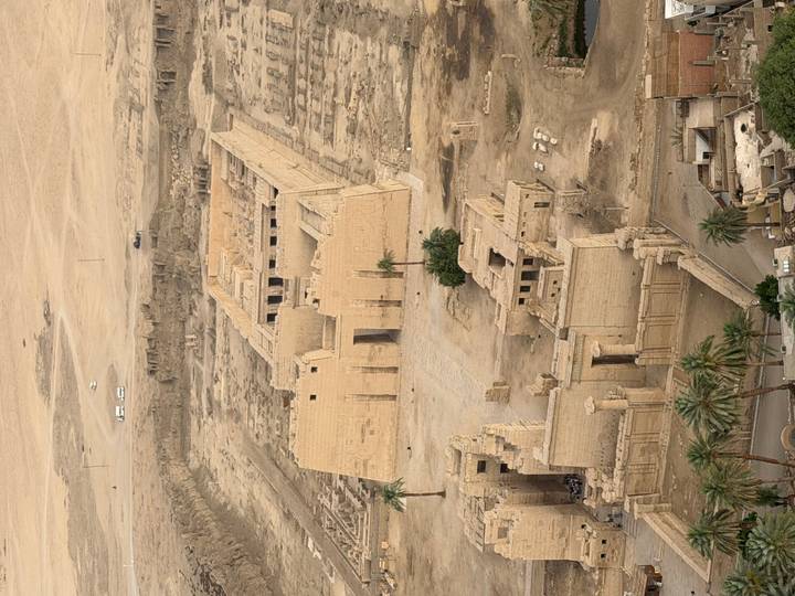 Aerial view of an ancient sandstone temple complex and surrounding desert landscape with palm trees at the edge.