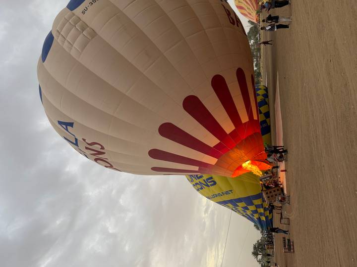 Ground crew inflates a colorful hot-air balloon at dawn, bright flame lighting the striped envelope against a cloudy sky.