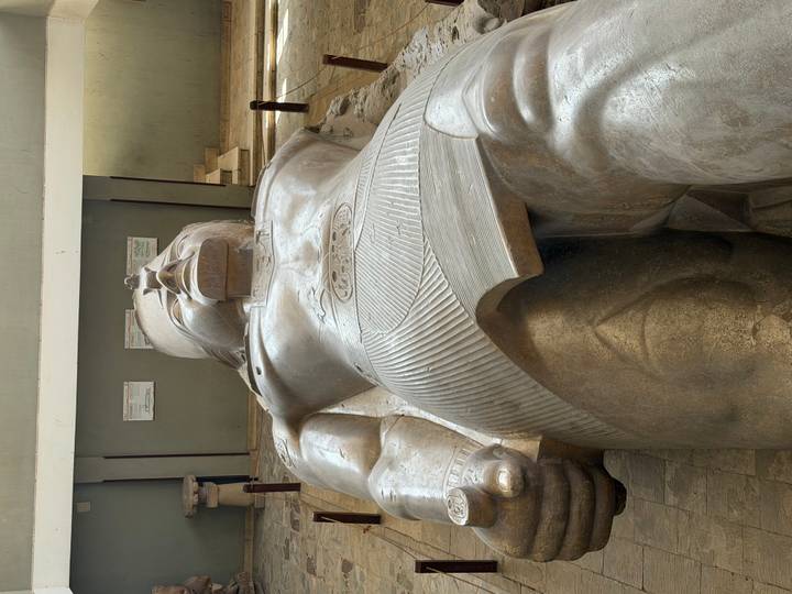 Massive recumbent statue of Pharaoh Ramesses II displayed indoors on a raised platform, viewed from the feet upward.
