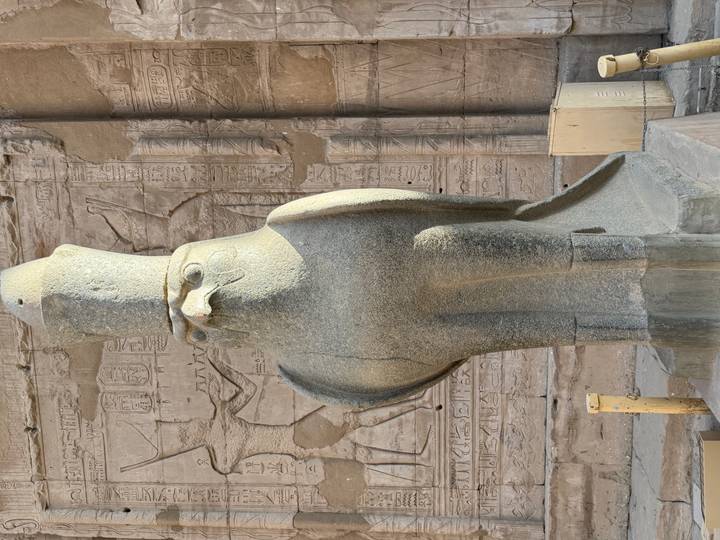 Stone statue of the falcon god Horus wearing a double crown in front of hieroglyphic-covered walls.
