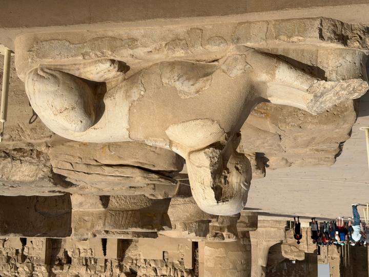 Weathered sandstone sphinx statue rests among temple ruins under bright sunlight.