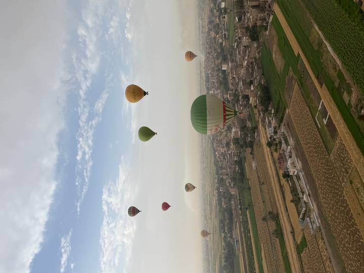Dozens of colorful hot-air balloons float over patchwork fields and small villages viewed from above.