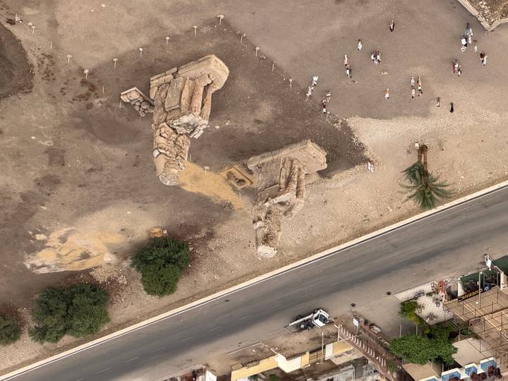 Aerial view of the Colossi of Memnon statues beside a road with small tourists gathered below.
