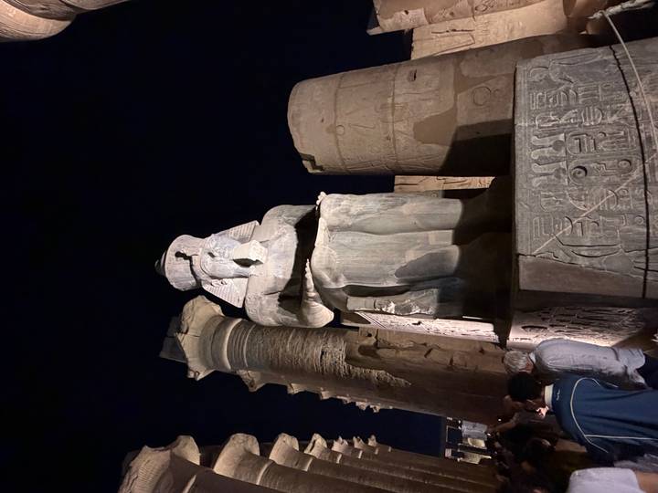 Night-lit statue seated among towering columns and hieroglyphic walls with visitors nearby.