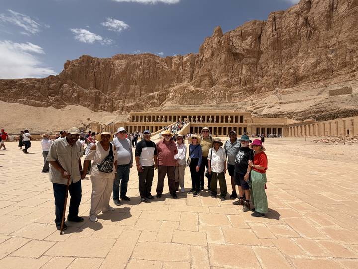 Tour group poses in front of Hatshepsut Temple nestled against dramatic cliffs under a bright sky.