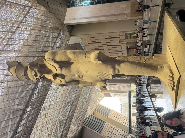 Grand indoor atrium of the Grand Egyptian Museum featuring colossal standing statue of Ramesses II surrounded by visitors.