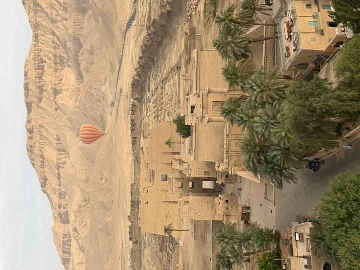 Hot-air balloon floats above temple ruins and lush palms with rugged desert hills behind.
